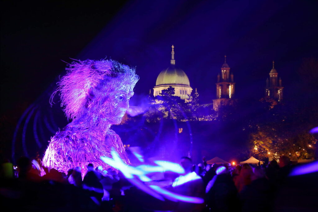 A large puppet of a giant's face is carried on the shoulders of people. There is a blue glow of nightlite. The procession is in the Macnas Parade for Galway Halloween